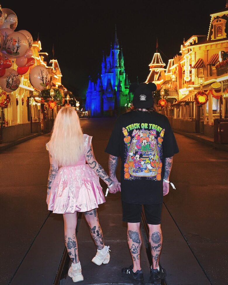 A couple walking hand in hand toward a lit castle at night, his black Mouse House tee printed large across the back
